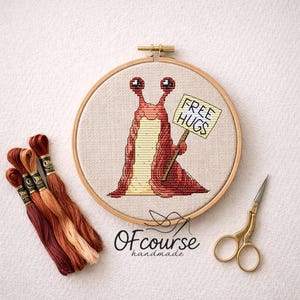 May include: A cross-stitch embroidery hoop with a cartoon slug holding a "FREE HUGS" sign. The slug is red and yellow, with large eyes. Embroidery floss in brown and gold shades are next to the hoop, along with gold scissors. The text "Ofcourse handmade" is at the bottom.