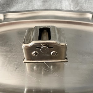 Peut inclure: Petit objet rectangulaire en métal argenté avec une fente sur le dessus et deux petits détails circulaires sur le devant. L'objet repose sur un plateau argenté, avec un tissu gris en arrière-plan.