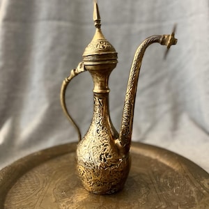 Könnte beinhalten: Eine antike Messingkanne mit kunstvollem Design, einem langen, gebogenen Ausguss und einem kleinen Griff. Sie steht auf einem passenden Messingtablett mit geätzten Mustern. Das Design deutet auf einen orientalischen oder Vintage-Stil hin. Höhe ca. 20 cm.