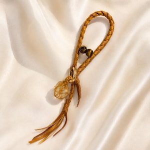May include: A braided tan leather strap keychain featuring two dark brown tiger eye beads, a gold-toned metal ring, and a clear, faceted crystal stone encased in a gold wire mesh cage, finished with a dangling leather fringe tassel at the end.