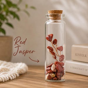 May include: A clear glass bottle filled with red jasper stones and a copper wire sculpture. The bottle is topped with a cork stopper. The words "Red Jasper" are written in a cursive font on the left side of the image.