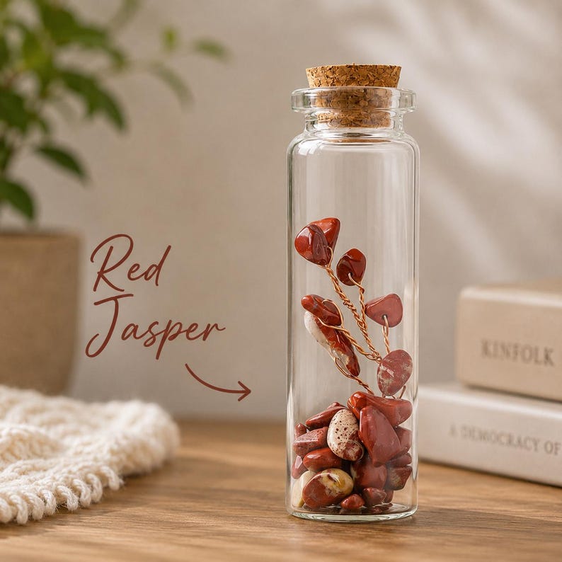 May include: A clear glass bottle filled with red jasper stones and a copper wire sculpture. The bottle is topped with a cork stopper. The words "Red Jasper" are written in a cursive font on the left side of the image.
