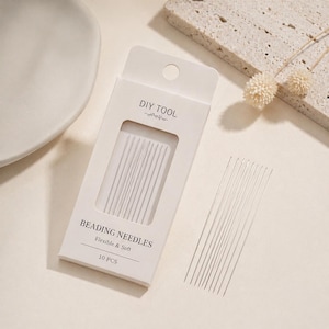 May include: A white box of beading needles with the text "DIY TOOL" and "BEADING NEEDLES Flexible & Soft 10 PCS". The box has a clear window showing the needles. Several needles are scattered outside the box.