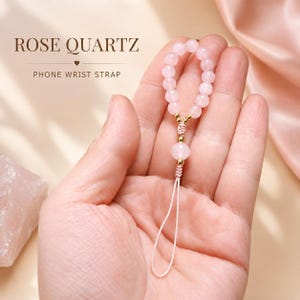 Puede incluir: Una correa de muñeca para teléfono con un bucle de cuentas de cuarzo rosa pálido acentuado con pequeñas esferas de metal dorado. El texto ROSE QUARTZ PHONE WRIST STRAP es visible. La correa incluye un cordón blanco fino para el dispositivo.