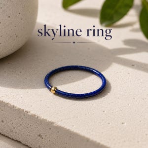 Op de afbeelding: Een dunne ring bestaande uit kleine, cilindrische diepblauwe kralen in een doorlopende cirkel, geaccentueerd door een enkele gladde, gepolijste goudkleurige bolvormige kraal. De tekst "skyline ring" staat boven het sieraad.