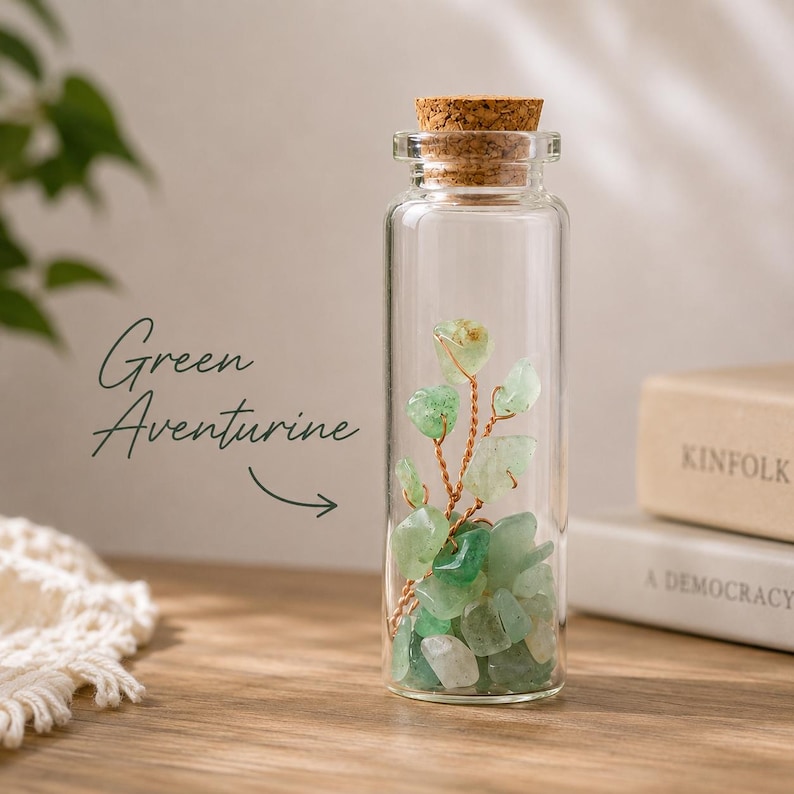 May include: A clear glass bottle with a cork stopper, filled with green aventurine stones and a wire sculpture. The text "Green Aventurine" is written on the left side of the image. The bottle is on a wooden surface.