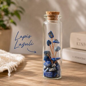 May include: A clear glass bottle filled with blue lapis lazuli stones and a copper wire tree. The bottle is topped with a cork stopper. The words "Lapis Lazuli" are written in blue script.