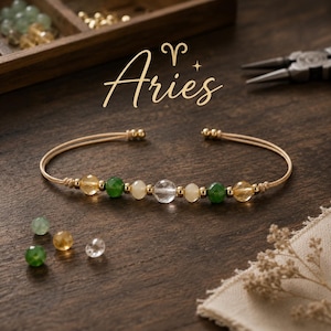 May include: Aries zodiac sign bracelet. The gold-coloured bracelet features a series of colourful beads, including green, yellow, and clear, strung along a beige cord. The word "Aries" is written in gold script above the bracelet.