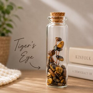 May include: A clear glass bottle with a cork stopper filled with tiger's eye stones. The stones are various shades of brown and gold. The text "Tiger's Eye" is written on the left side of the image.