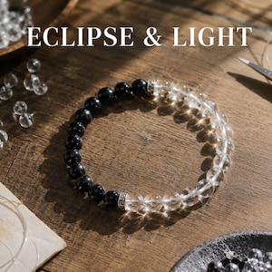 May include: A beaded bracelet featuring alternating sections of polished black beads and clear, translucent beads. The bracelet includes two silver-toned metal spacer beads with intricate detailing. The text ECLIPSE & LIGHT is visible.