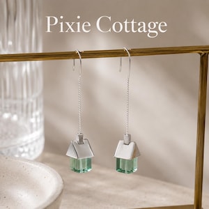 Puede incluir: Un par de pendientes colgantes plateados con forma de casa, con un techo metálico y una base cúbica de color verde translúcido. Los pendientes cuelgan de finas cadenas y ganchos plateados. El texto Pixie Cottage es visible sobre ellos.