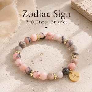 Könnte beinhalten: Ein rosa Kristallarmband mit einem goldenen Anhänger. Das Armband hat rosa und graue Perlen, die durch goldfarbene Abstandshalter mit klaren Kristallen getrennt sind. Der Anhänger hat eine goldene Oberfläche und ein Tierkreiszeichen. Der Text auf dem Bild lautet "Zodiac Sign Pink Crystal Bracelet."