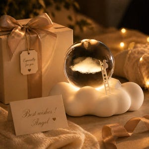 May include: A decorative snow globe with a cloud scene and a tiny figure on a ladder. The globe is set on a white cloud-shaped base that glows softly. A gift box with a bow and a card reading "Best wishes! Angel" are also visible.