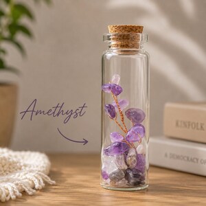May include: A clear glass bottle filled with amethyst crystals and a cork stopper. The word "Amethyst" is written in a cursive font. The amethyst crystals are various shades of purple and white.