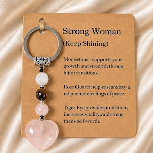 May include: A silver-toned metal keychain featuring a round ring and a vertical strand of four beads: a white moonstone, a dark brown tiger eye, a light pink rose quartz, and a large heart-shaped rose quartz pendant attached to a tan card.