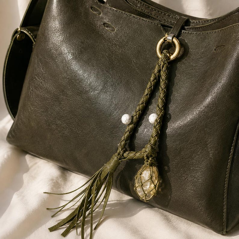 May include: A dark olive green leather shoulder bag featuring a decorative accessory attached to a gold-toned metal ring. The accessory consists of two braided olive green cords, two small white stone beads, and a clear crystal encased in netting.