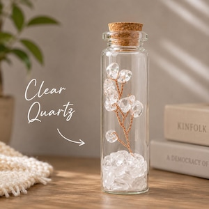 May include: A clear glass bottle with a cork stopper, filled with clear quartz crystals. Inside, a copper wire sculpture with quartz crystal accents resembles a small tree. The words "Clear Quartz" are written on the left.