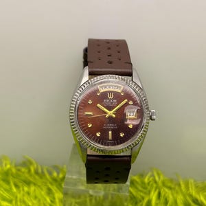 May include: A vintage Ricoh automatic watch with a brown dial and a silver-tone case. The watch features a brown leather strap and displays the day of the week and date. The watch face has gold-tone hands and markers. The watch is labeled "21 Jewels Shockproof".