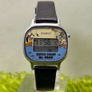 May include: Vintage Casio digital watch with a black leather strap. The watch face features a cartoon scene with the text "Quick Draw Mc Graw" and the time 11:56. The watch is silver-toned with a black strap and the Casio logo.