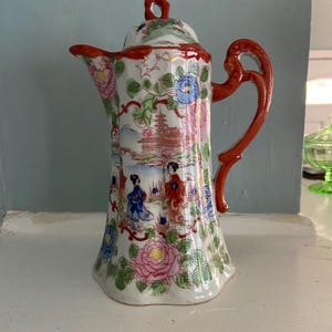 May include: A white porcelain teapot with a red handle and spout. The teapot is decorated with a floral and landscape design in pink, blue, and green. The lid has a red trim and a small loop handle.