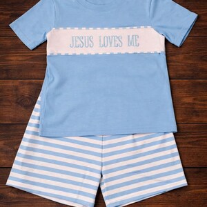 May include: A light blue two-piece pajama set. The short-sleeved top has a white band across the chest with the text "JESUS LOVES ME". The matching shorts are light blue and white horizontal stripes. The set is made of soft fabric.