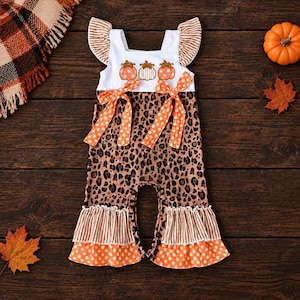 May include: A sleeveless romper featuring a white bodice with three pumpkin graphics, ruffled shoulders, and two orange polka-dot waist ties. The lower portion has a brown leopard print pattern, finished with striped and orange polka-dot ruffled hems.
