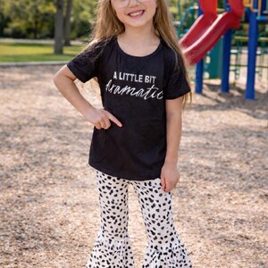 May include: A young girl wearing a black t-shirt with the text "A LITTLE BIT dramatic" in white script. She is also wearing white pants with black spots and flared bottoms. The girl is smiling and pointing at the shirt.