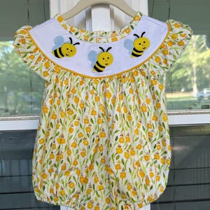 May include: A baby romper with a white collar and yellow trim, adorned with three embroidered bees. The romper features a floral pattern with yellow flowers and green leaves on a white background. The garment is hanging on a beige hanger.