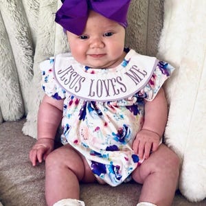 May include: A baby wearing a floral print romper with the text "JESUS LOVES ME" embroidered on the collar. The baby is also wearing a purple bow headband and white lace-trimmed socks. The romper features a watercolor floral design with blue, purple, and pink hues.