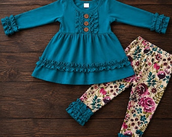 May include: A teal long-sleeve tunic featuring a ruffled bodice with three brown buttons, a ruffled hem, and ruffled sleeve cuffs. The outfit includes matching leggings with a cream background and a floral print of roses and leaves.