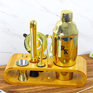 May include: A gold-colored cocktail shaker set with various bar tools, including a shaker, strainer, muddler, jigger, and bar spoon. The set is displayed on a bamboo tray. The shaker has the initials "RK" on it.