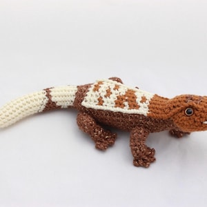 May include: A crocheted lizard toy in shades of brown, cream, and white. The lizard features a patterned back and tail, with small, detailed legs and a smiling face. The toy is approximately 10 inches long.