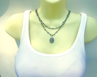 May include: A layered necklace with a silver pendant featuring a detailed design. The necklace is made of dark grey metal beads and chains.