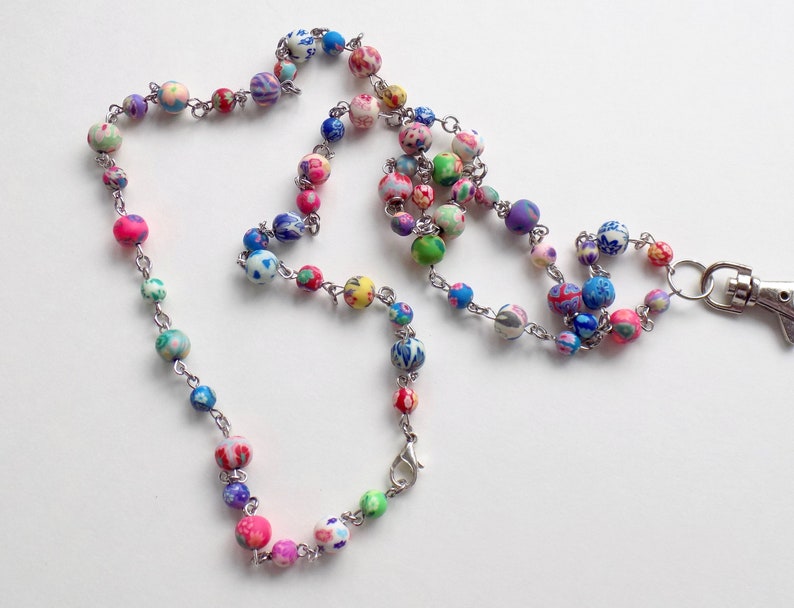 May include: A colorful beaded lanyard with a silver clasp. The beads are made of polymer clay and feature a variety of floral patterns in shades of pink, blue, green, and white.