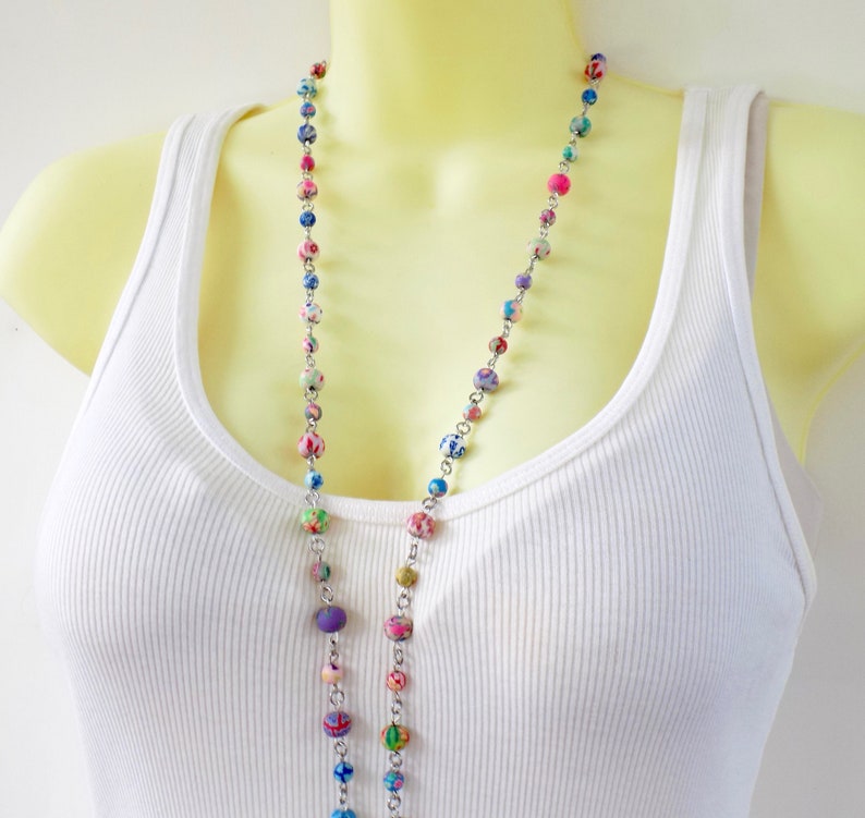 May include: A long silver chain necklace with colorful patterned beads. The beads are a mix of purple, blue, green, pink, and white.