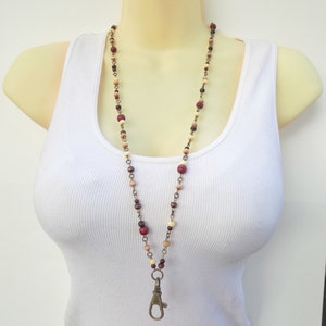 May include: A long beaded lanyard necklace with a lobster clasp. The necklace features wooden beads in red, brown, and cream, strung with a metal chain. The clasp is bronze-coloured.