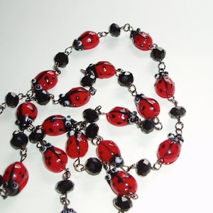 May include: A necklace featuring a series of red and black ladybug-shaped beads interspersed with faceted black beads. The beads are connected by thin metal links, creating a continuous chain design with a repeating pattern of insects and dark stones.