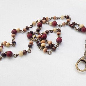 May include: A beaded lanyard with wooden beads in red, brown, and cream. The beads are strung on a bronze-coloured chain and finished with a bronze-coloured lobster clasp.