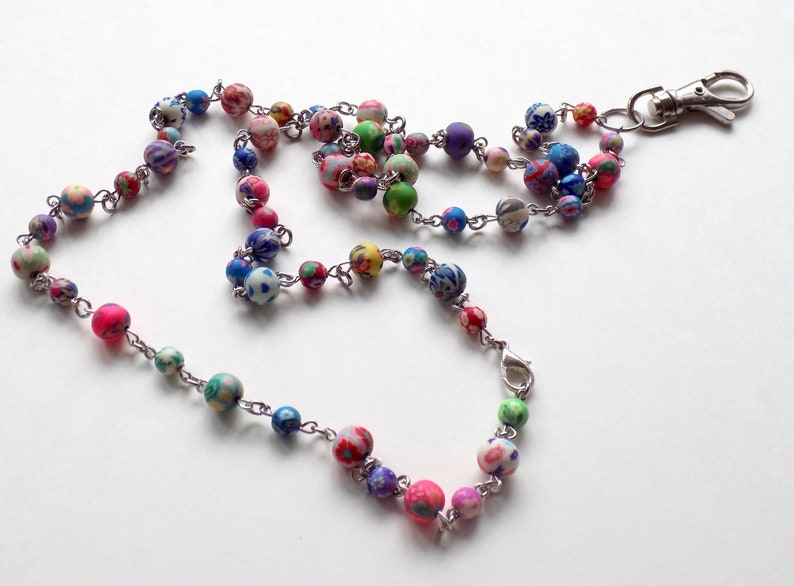 May include: A colorful beaded lanyard with a silver chain and a silver clip. The beads are made of polymer clay and have a variety of patterns and colors, including pink, blue, green, and white.