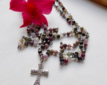 May include: A silver cross pendant with a clear crystal hangs from a multi-colored beaded necklace. The beads are pink, green, black, and white.