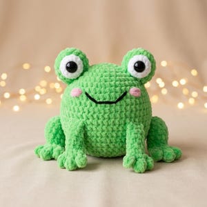 May include: A handmade green crochet frog with large white and black eyes, pink cheeks, and a smiling mouth. The frog has detailed feet and is a plush toy.