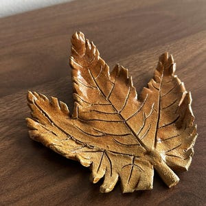 May include: A detailed, golden-brown leaf-shaped decorative object. The leaf features intricate vein detailing and a textured surface. The object is likely made of metal or a similar material, and it is designed to be a decorative accent.