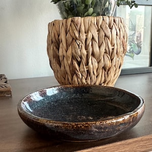 Op de afbeelding: Een donkerblauwe en bruine keramische kom met een ondiepe, afgeronde vorm. De kom heeft een glanzende afwerking en staat op een houten oppervlak. Een gevlochten mand en een glazen kom met groen staan op de achtergrond.