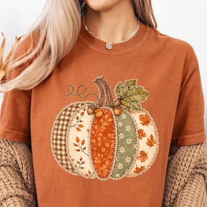 May include: A rust-coloured short-sleeve t-shirt featuring a large central graphic of a pumpkin. The pumpkin is composed of various patchwork-style segments in shades of brown, orange, and green, decorated with patterns of leaves, flowers, and gingham.