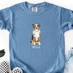 May include: A blue short-sleeve t-shirt featuring a graphic of a sitting Australian Shepherd dog with the name BELLA printed in white underneath. The shirt is displayed with black sunglasses, a pair of black and white checkered slip-on shoes, and light blue denim shorts.
