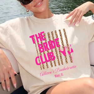 May include: A beige t-shirt with the text "THE BRIDE CLUB" in pink and leopard print stripes. The text "Allison's Bachelorette Miami, FL" is printed below. The shirt is worn by a person wearing sunglasses.