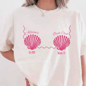 以下が含まれることがあります： ピンクの貝殻のデザインが施されたクリーム色のTシャツ。「Allison's Bach Club」の文字と「Est 2026」と「Miami, FL」の文字がプリントされています。赤と白のチェック柄のショートパンツを着用しています。