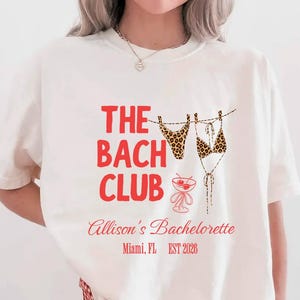 May include: A cream-coloured t-shirt with the red text "THE BACH CLUB", "Allison's Bachelorette, Miami, FL, EST 2026", a leopard print bikini graphic and a cocktail illustration. The model is wearing red and white checkered shorts.