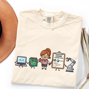 以下が含まれることがあります： クリーム色のTシャツ。リンゴを持った笑顔のキャラクター、ノートパソコン、回路基板、CODING LESSON PLANと書かれたクリップボード、プログラミングアイコン、ロボットアームが描かれたグラフィックがプリントされています。