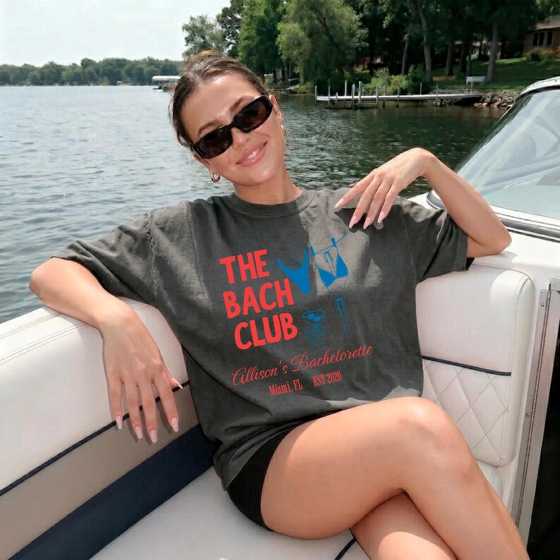 May include: Dark grey t-shirt with the text "THE BACH CLUB" in red and a blue bikini graphic. The shirt also includes the text "Allison's Bachelorette Miami, FL EST 2023". The person is wearing black sunglasses and black shorts.